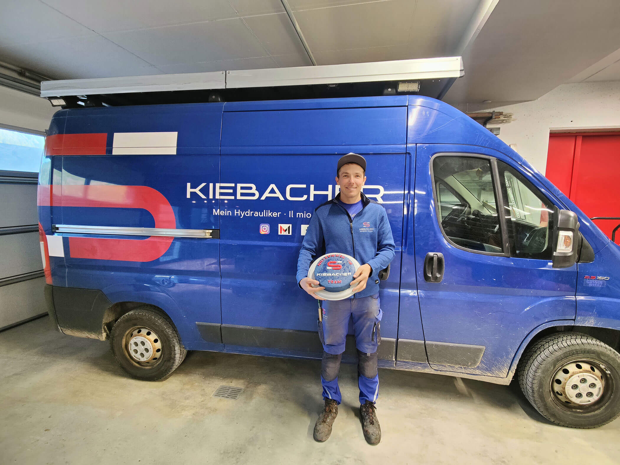 Simon Kiebacher davanti alla sua auto aziendale con una torta in mano - Kiebacher Wahlen